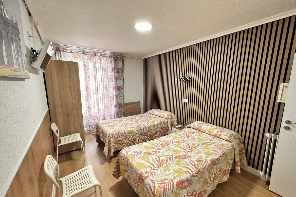 Una habitación con dos camas, una silla, un armario de madera, un radiador y una lámpara fijada a la pared.