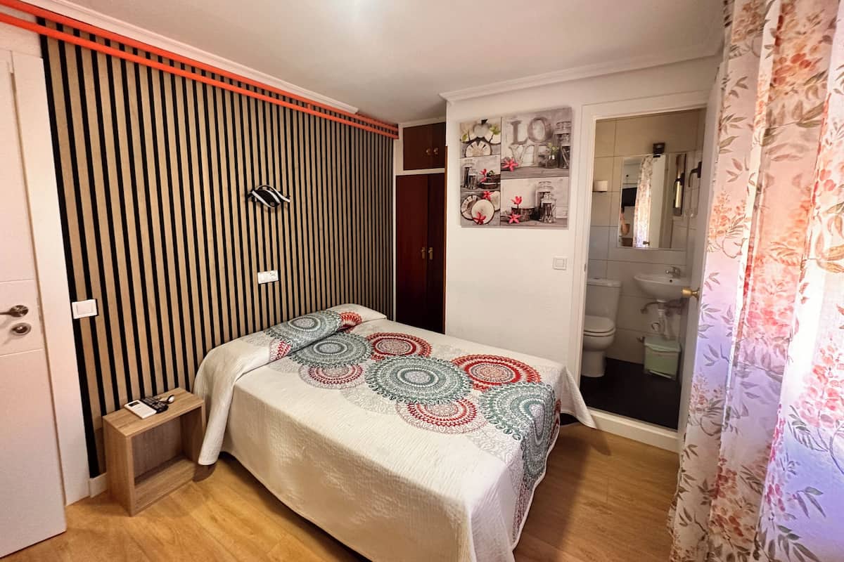 Un dormitorio con cama, una mesita de noche, estante de pared con objetos decorativos, una puerta y un baño visible a través de una puerta abierta.
