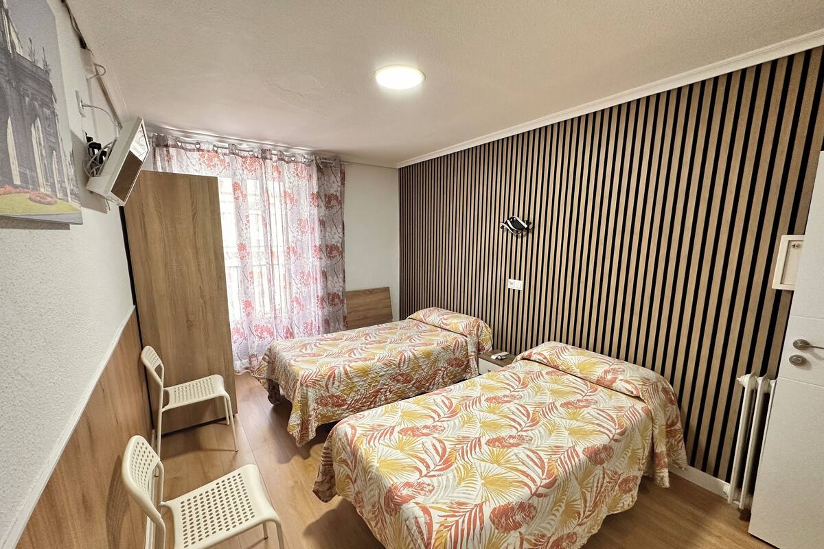 Una habitación con dos camas, una silla, un armario de madera, un radiador y una lámpara fijada a la pared.