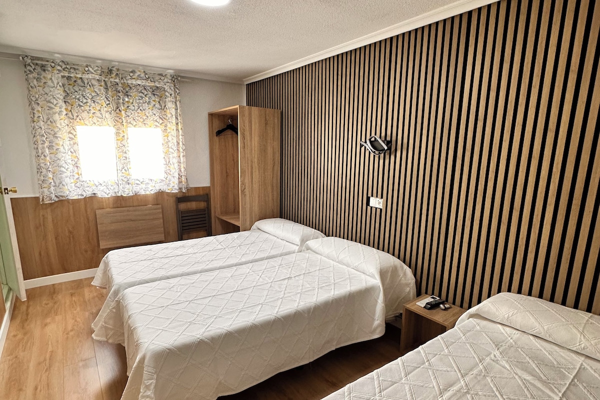 A hotel room with three beds, a wooden wardrobe, a small table with a lamp, and a window with patterned curtains.