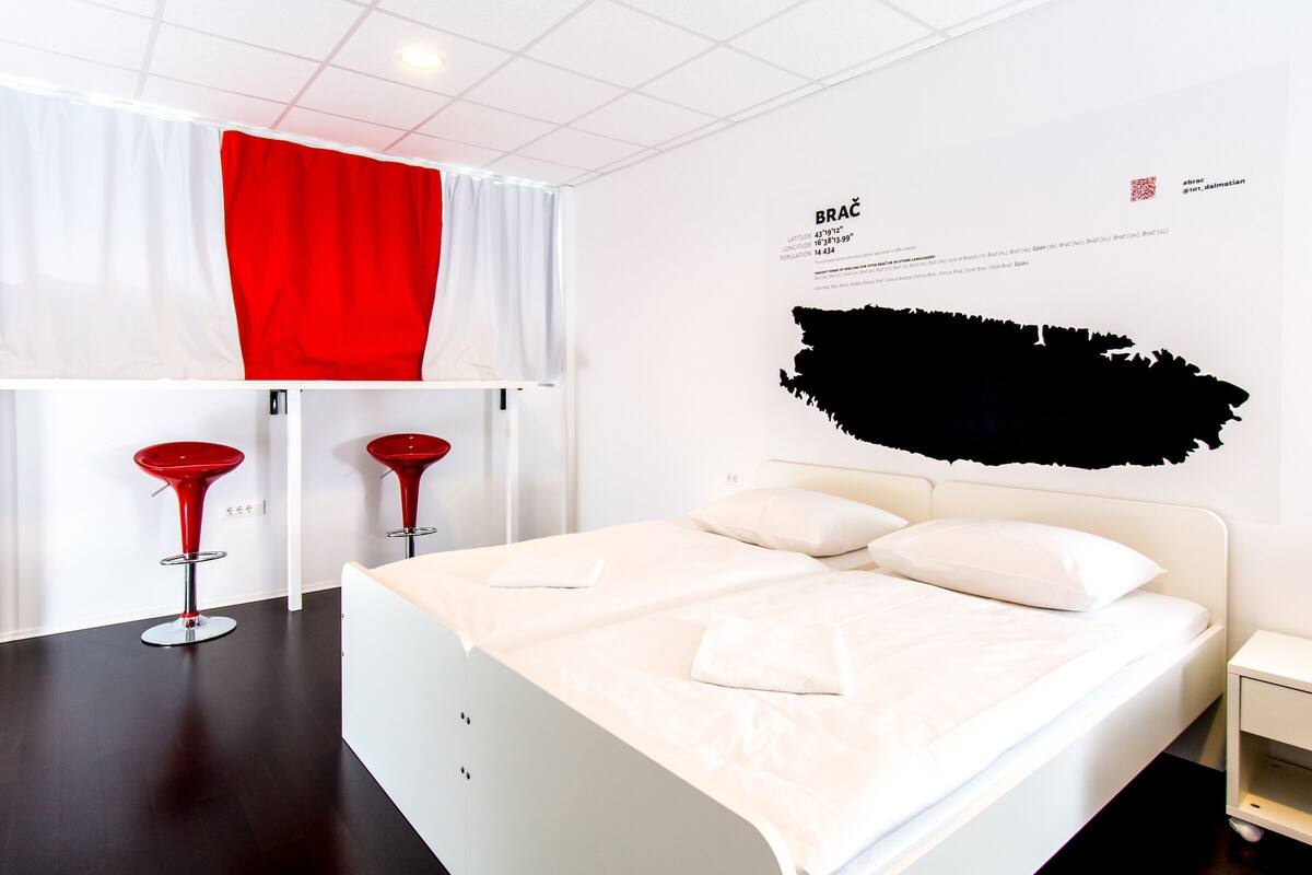 Ein modernes Hotelzimmer mit einem Bett, zwei roten Barhocker, einem weißen Vorhang und einer Wand mit Text und einer schwarzen Form.