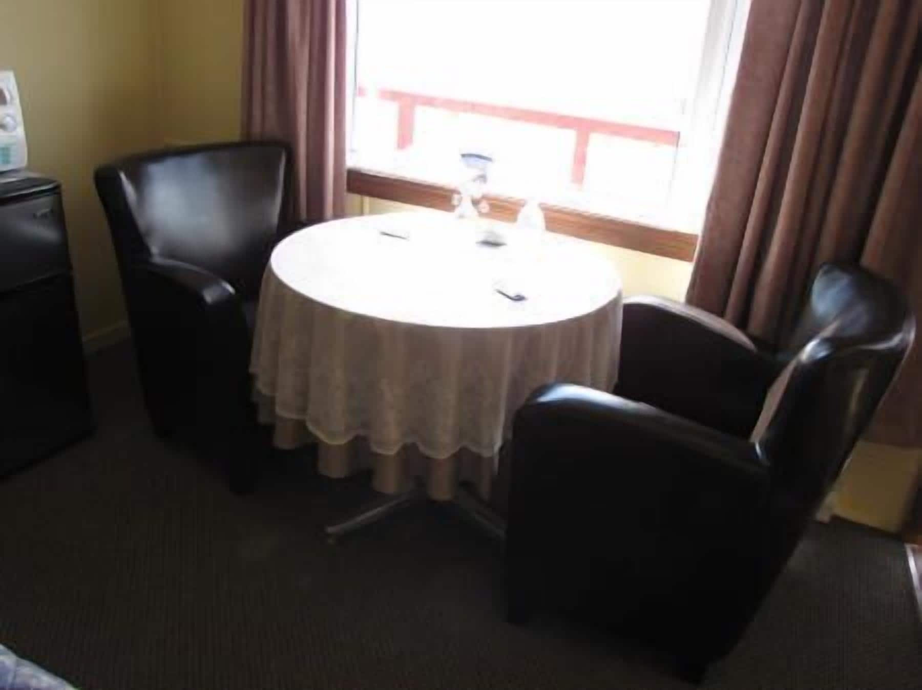 A small room with a round table and two chairs, a window with curtains, and a small desk with a lamp.