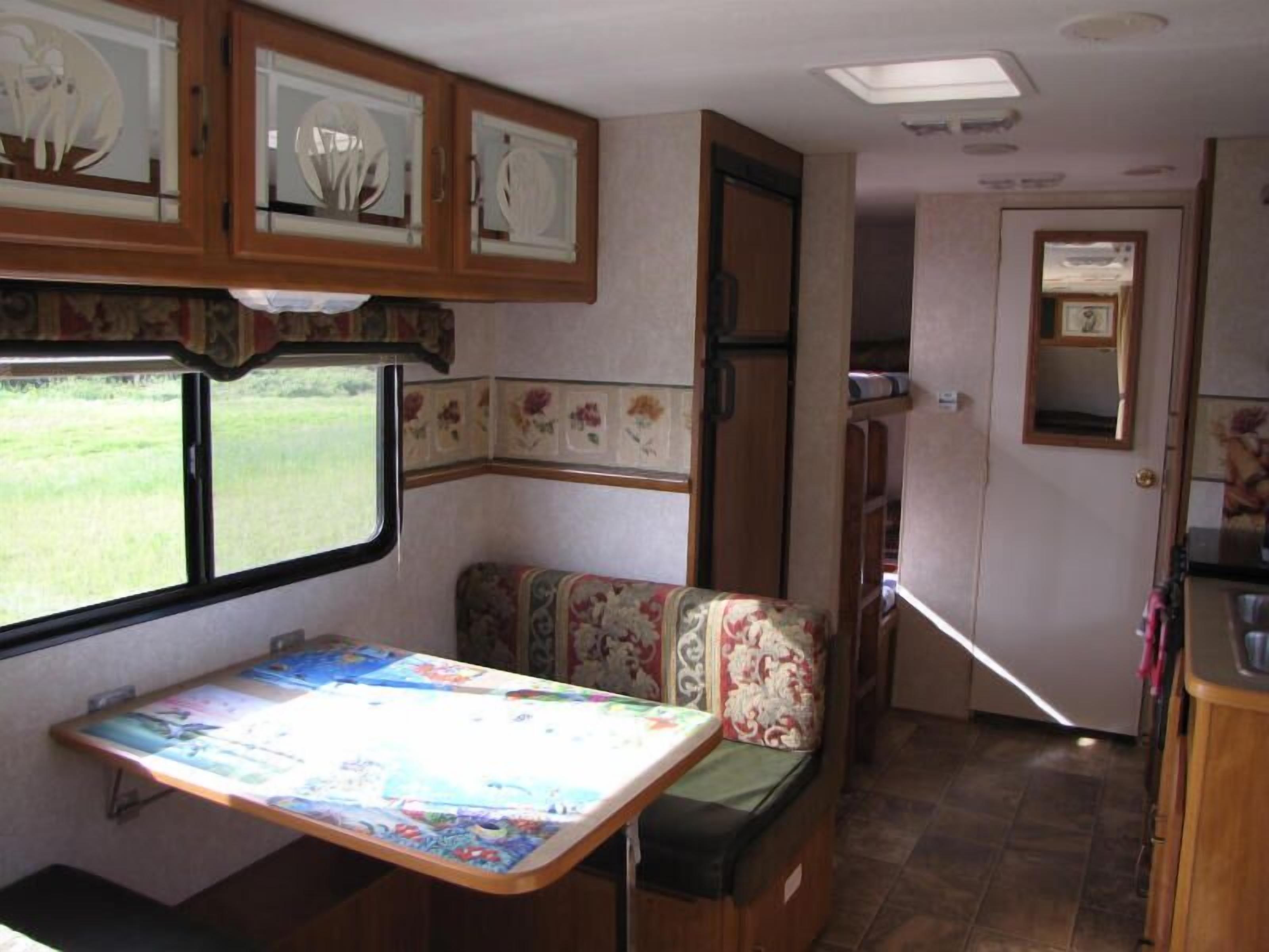 Interior compacto de RV com cozinha compacta, área de refeições com mesa e banco, e janela com vista para o exterior.