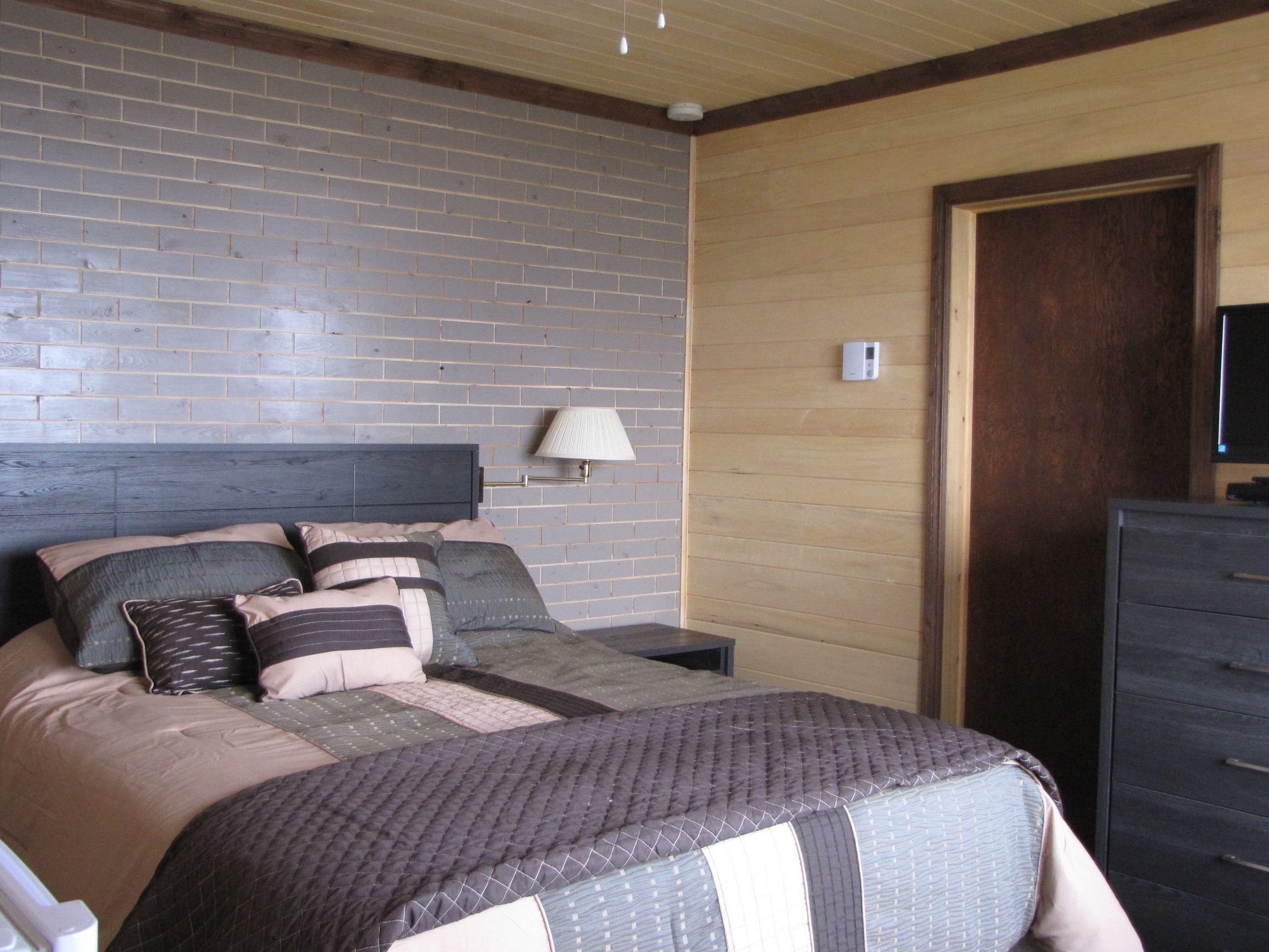 Une chambre à coucher avec une tête de lit ornée d’un motif en briques, un lit avec de nombreux oreillers, une lampe de chevet, une porte en bois et une commode.