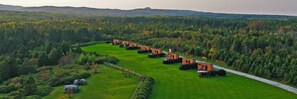 A series of small, rustic cabins arranged in a semi-circle on a lush green field, surrounded by dense forest. - Motel du Haut Phare (Gaspé)