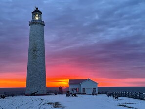 A lighthouse with a small adjacent building, both situated on a snowy landscape with a sunset in the background. - Motel du Haut Phare (Gaspé)