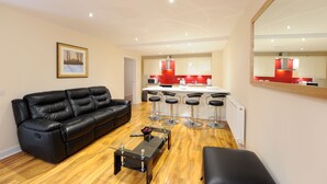 Una sala de estar moderna con un sofá de cuero negro, una mesa de centro de vidrio y una cocina con electrodomésticos rojos y taburetes de bar.