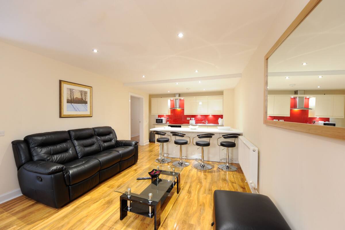 A modern living room with a black leather sofa, a glass coffee table, and a kitchen area with red appliances and bar stools.