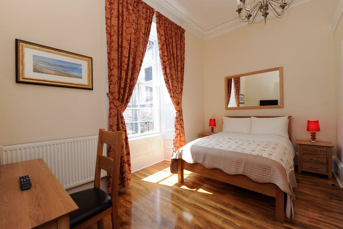 A bedroom with a bed, a bedside table with a lamp, a chair, a window with curtains, a radiator, and a framed picture on the wall.