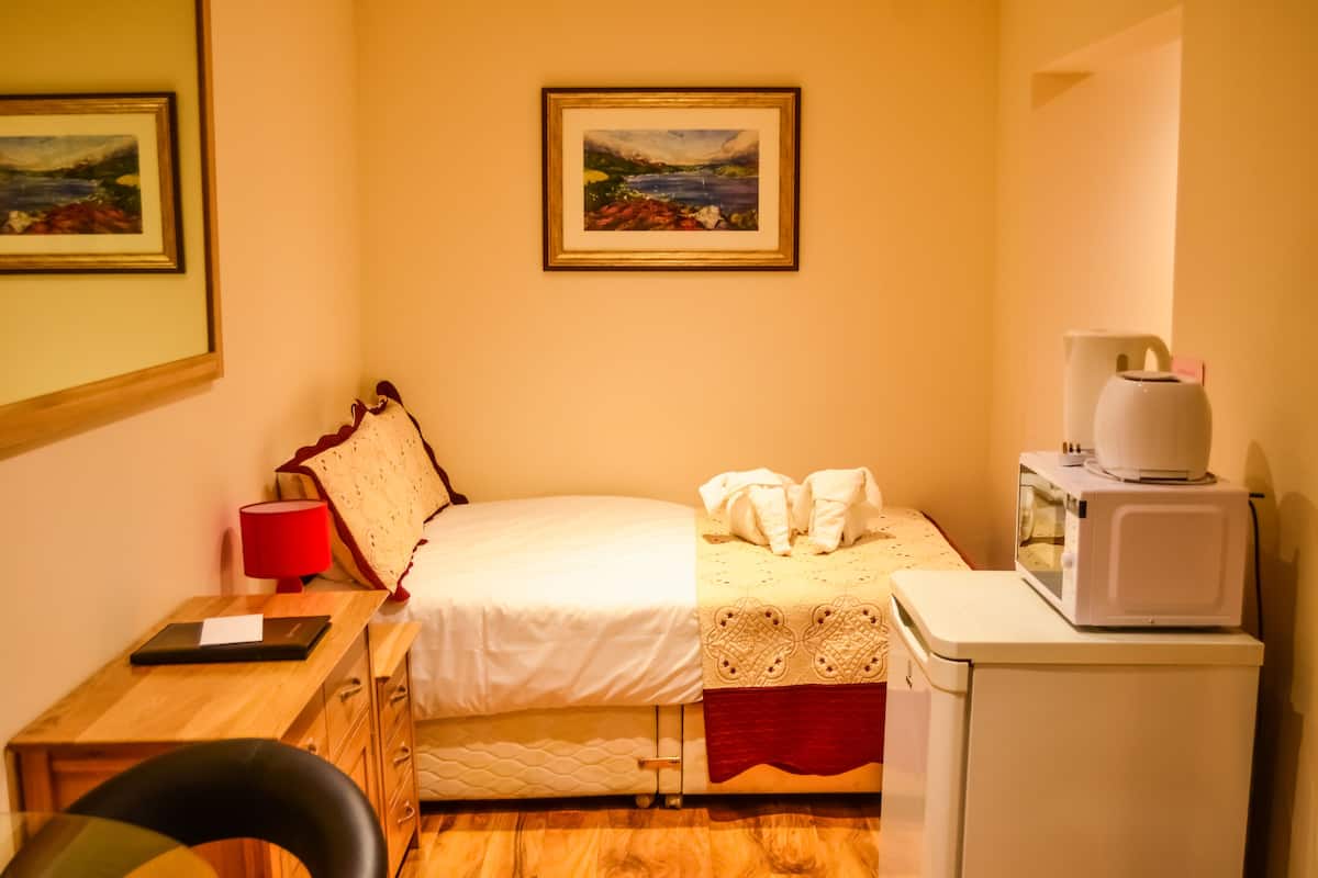 A small, cozy room with a bed, a desk, a chair, a microwave, and a framed painting on the wall.