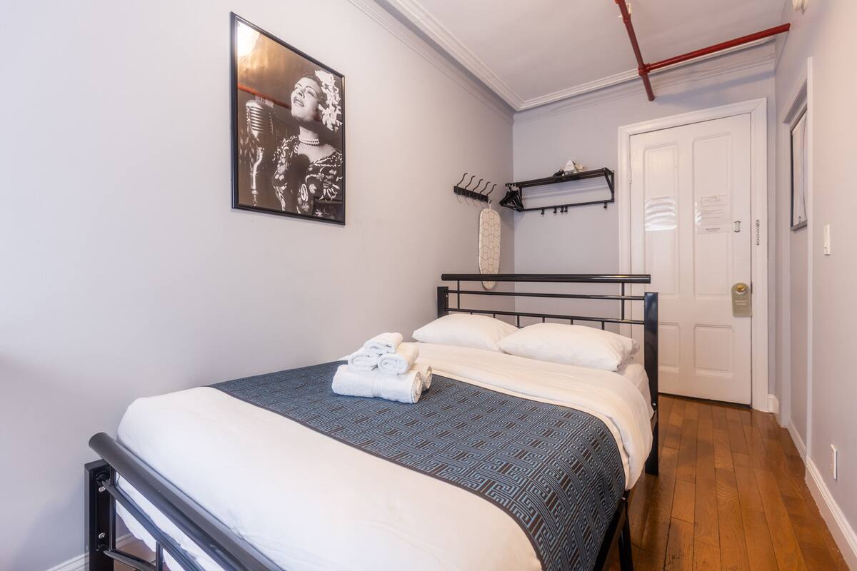 Un lit bien fait, avec une couvre-lit à motifs et des serviettes pliées, une tête de lit en métal noir, une étagère fixée au mur ornée d’un objet décoratif, et une porte avec une poignée.