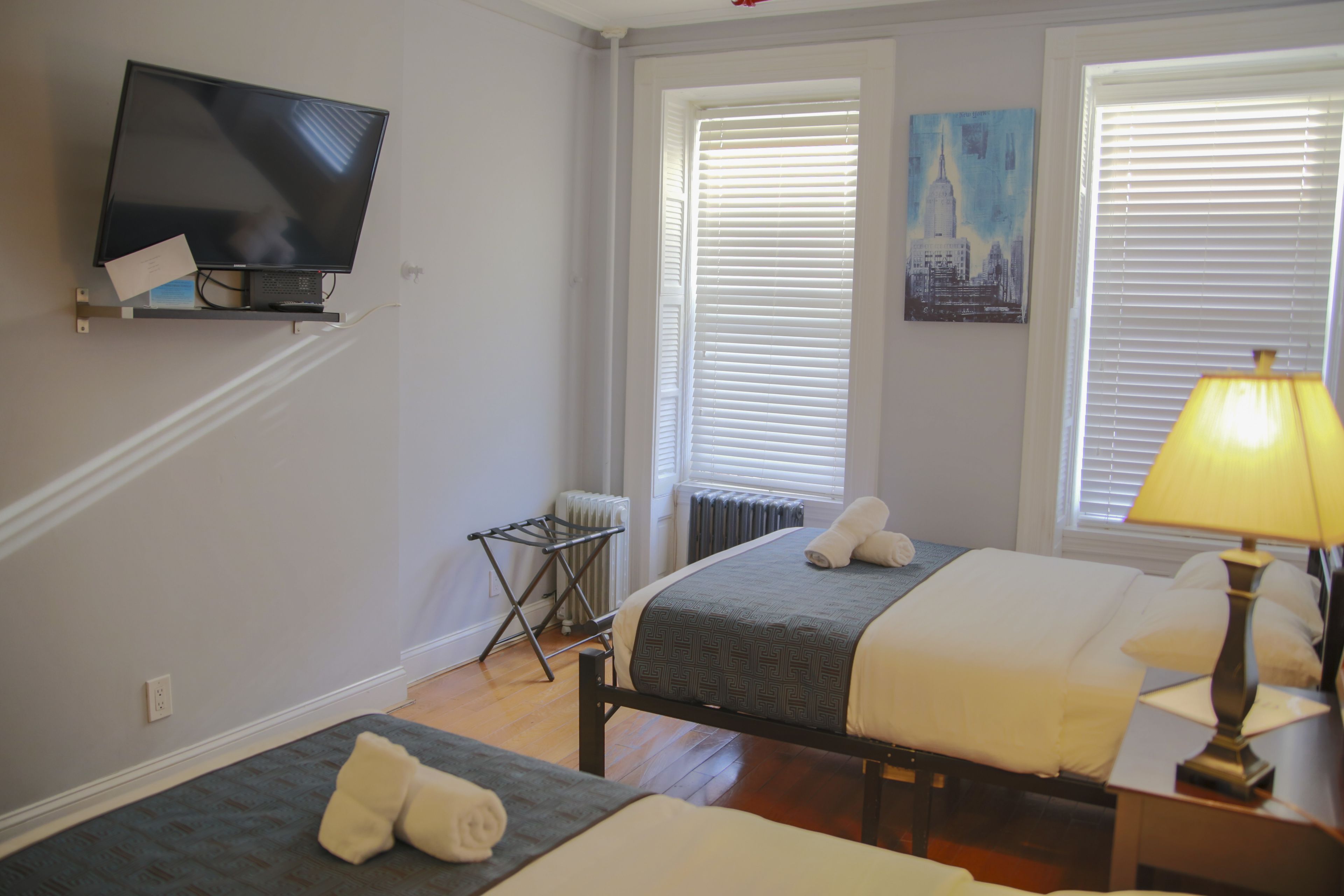A hotel room with two beds, a TV mounted on the wall, a radiator, and a window with blinds.