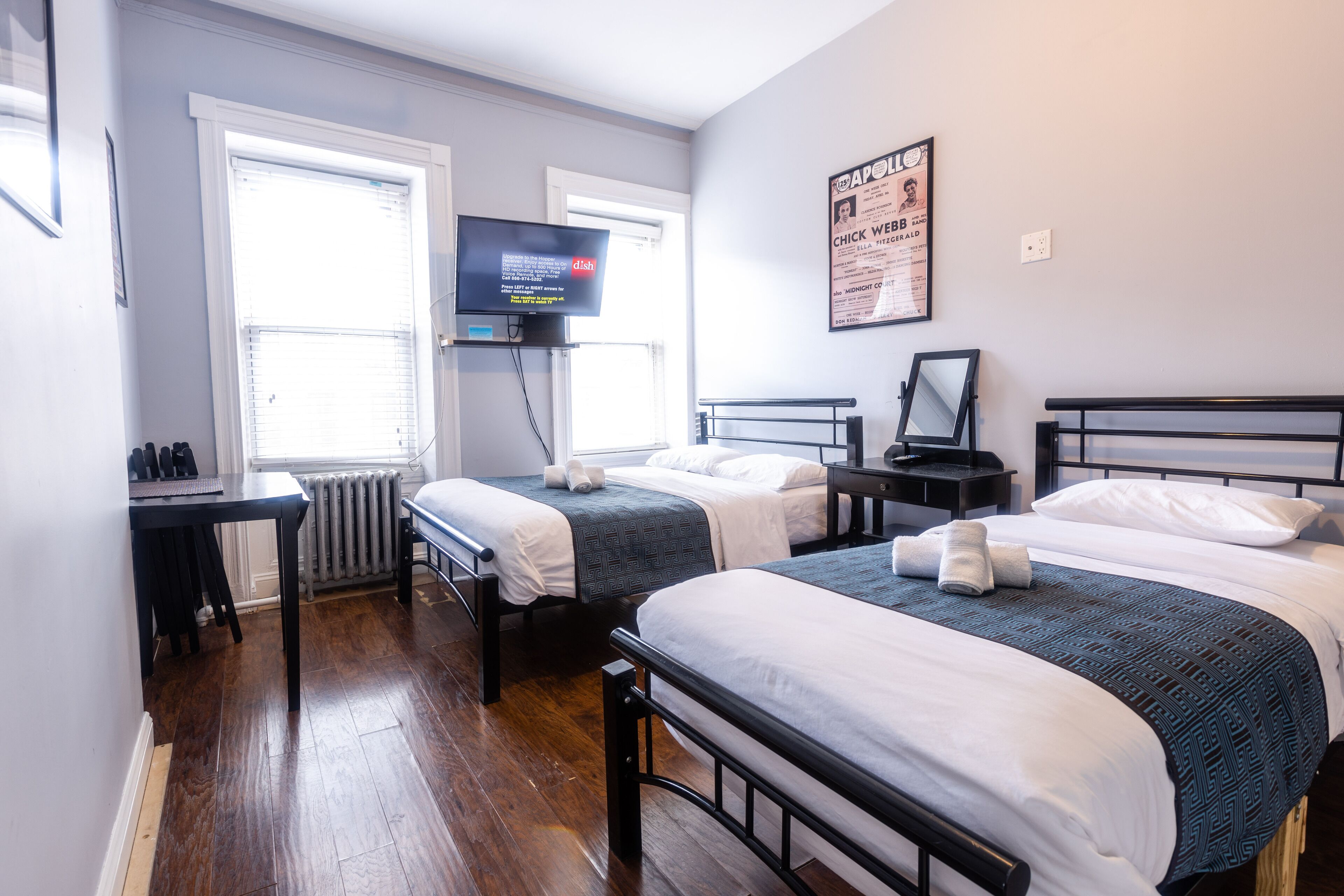 A hotel room with two single beds, a TV on the wall, a poster, and a window with blinds.