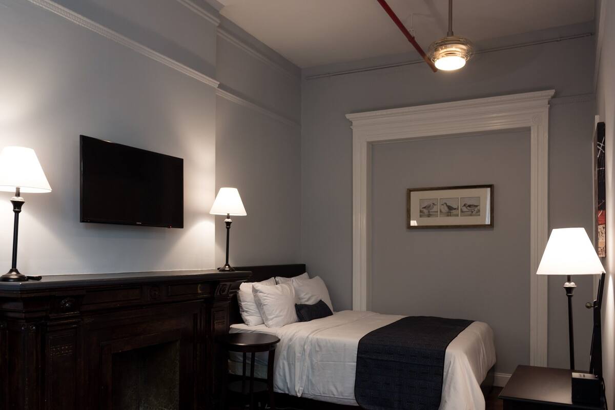 Une chambre d’hôtel avec un lit, deux lampes de chevet, un téléviseur à écran plat, une cheminée et un tableau encadré au mur.