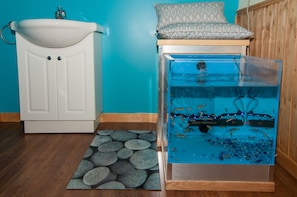 A bathroom with a white sink, a fish tank, and a patterned pillow on a wooden shelf.