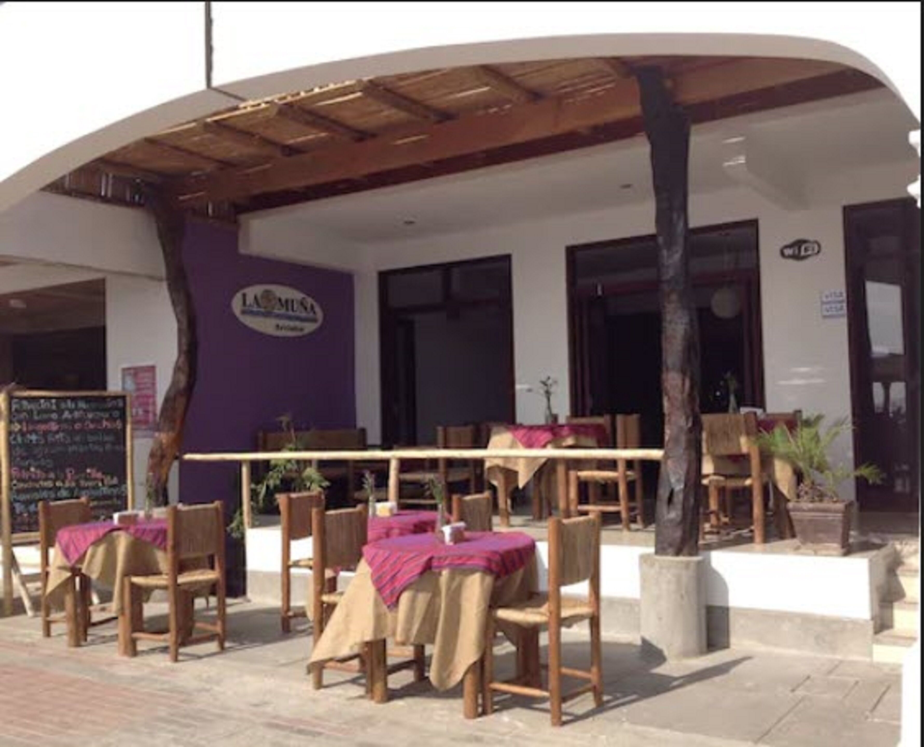 An outdoor seating area with wooden tables and chairs, covered by a wooden canopy. There is a sign on the wall and a chalkboard with writing.