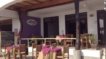 An outdoor seating area with wooden tables and chairs, covered by a wooden canopy. There is a sign on the wall and a chalkboard with writing.