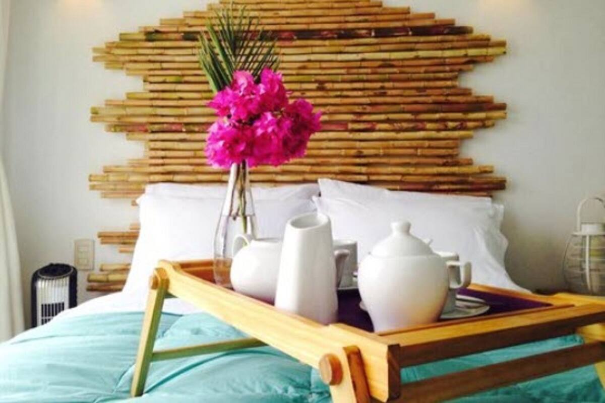 A neatly made bed with a wooden headboard, a tray with white ceramic items, and a vase of pink flowers.