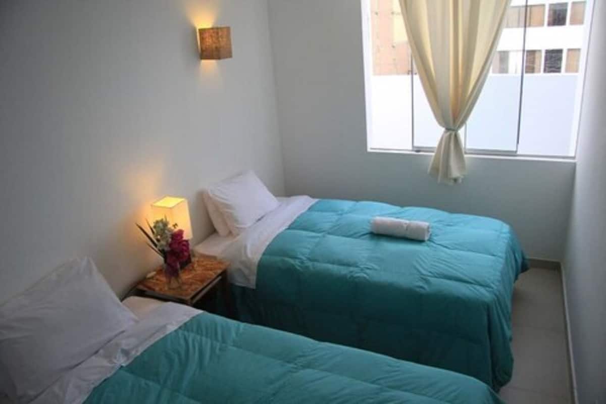 A room with two beds, a nightstand with a lamp, and a window with curtains.