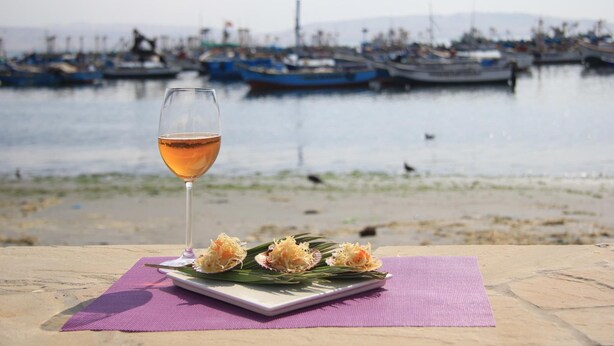 Una mesa con una copa de vino y un plato de comida frente a un muelle, con barcos al fondo.