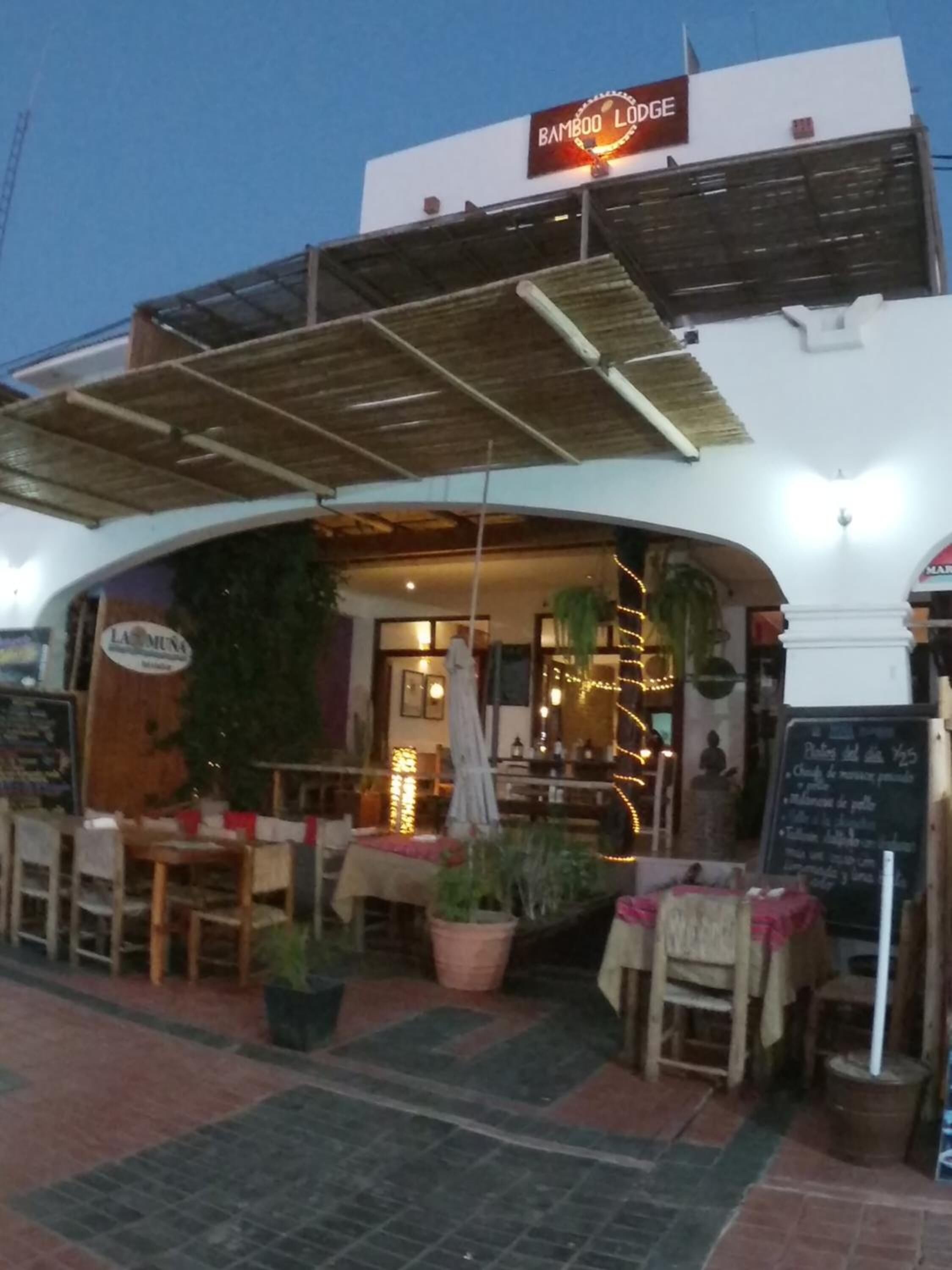 Un área de comedor al aire libre con mesas y sillas, un letrero que dice 'Bamboo Lodge' y una pizarra con el menú.
