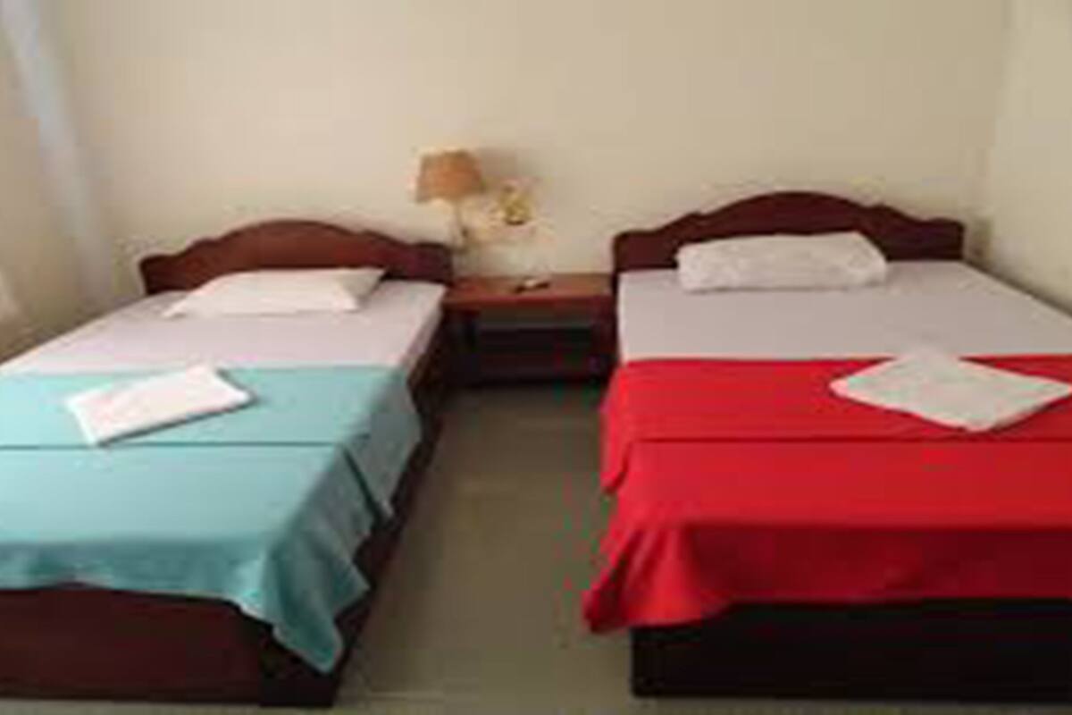 Une chambre avec deux lits simples, chacun recouvert d’une couvre-lit de couleur différente, une table de chevet avec une lampe et une fenêtre avec des rideaux.