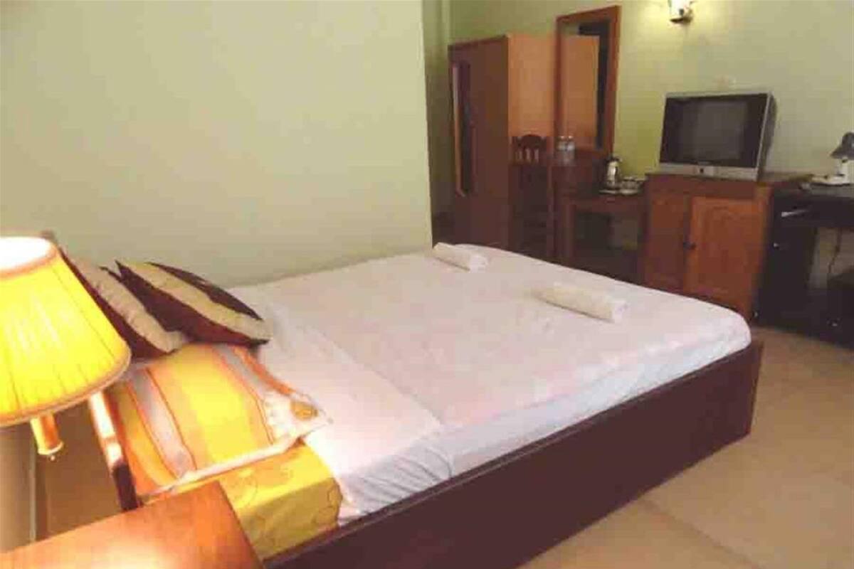 Un lit simple avec un cadre en bois, une table de chevet avec une lampe, un téléviseur posé sur un meuble et une armoire dans une pièce aux murs unis.