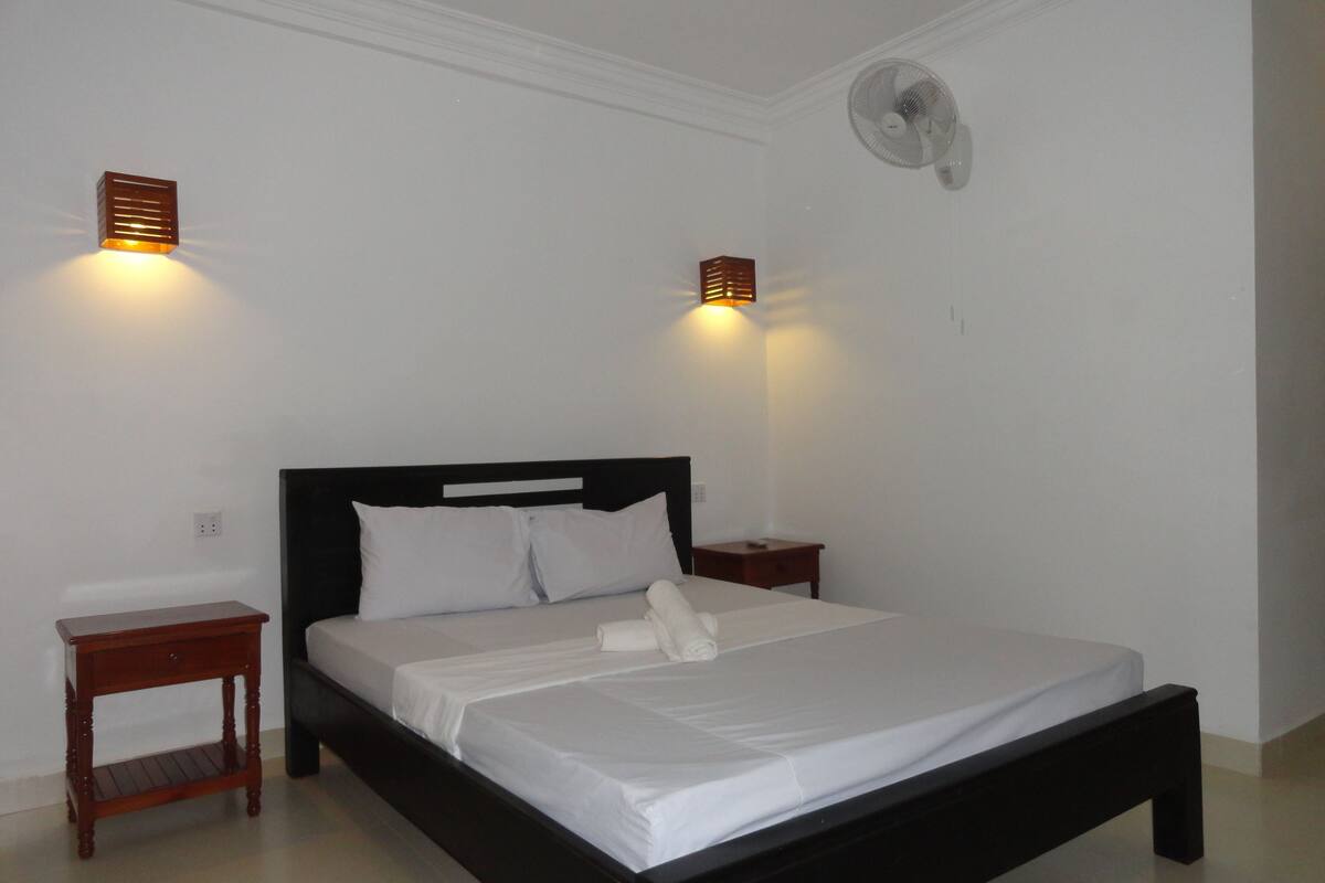Un lit bien fait, avec du linge de lit blanc et des oreillers, une table de chevet en bois avec une lampe, et un ventilateur fixé au mur.