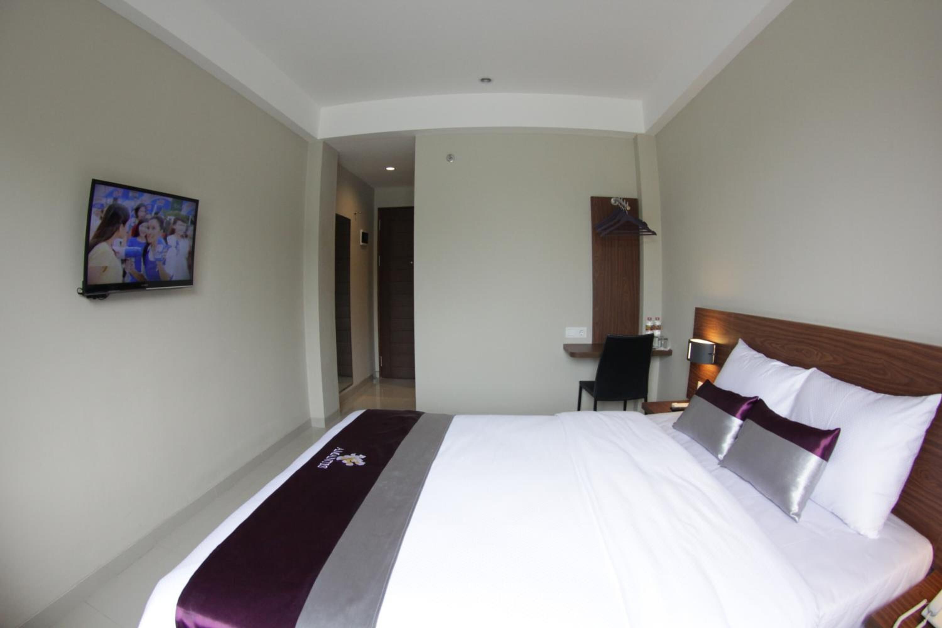 A hotel room with a large bed, a television mounted on the wall, a desk with a chair, and a wooden headboard.