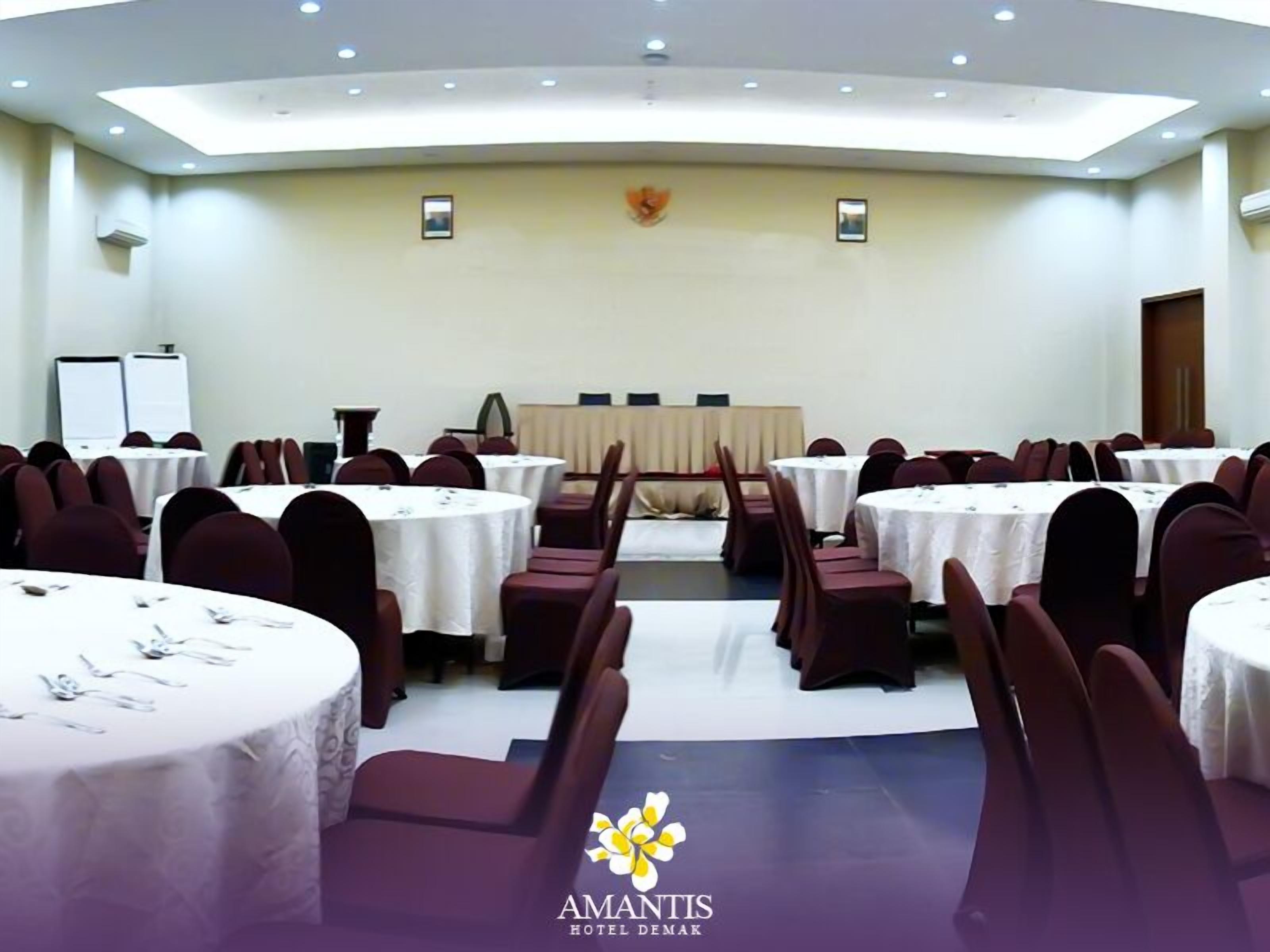 Uma sala de conferências com mesas e cadeiras redondas, um palco com um púlpito e o logotipo do 'Amantis Hotel Demak' na parte inferior.