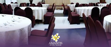 A conference room with round tables and chairs, a stage with a podium, and a logo for 'Amantis Hotel Demak' at the bottom.