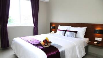 Cama bem arrumada com colcha roxa, cesta de frutas ao lado e mesa de cabeceira com abajur e telefone.