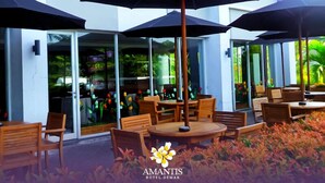 An outdoor seating area with wooden tables and chairs, large umbrellas, and a view of the hotel interior through glass doors. - Amantis Hotel (Demak)