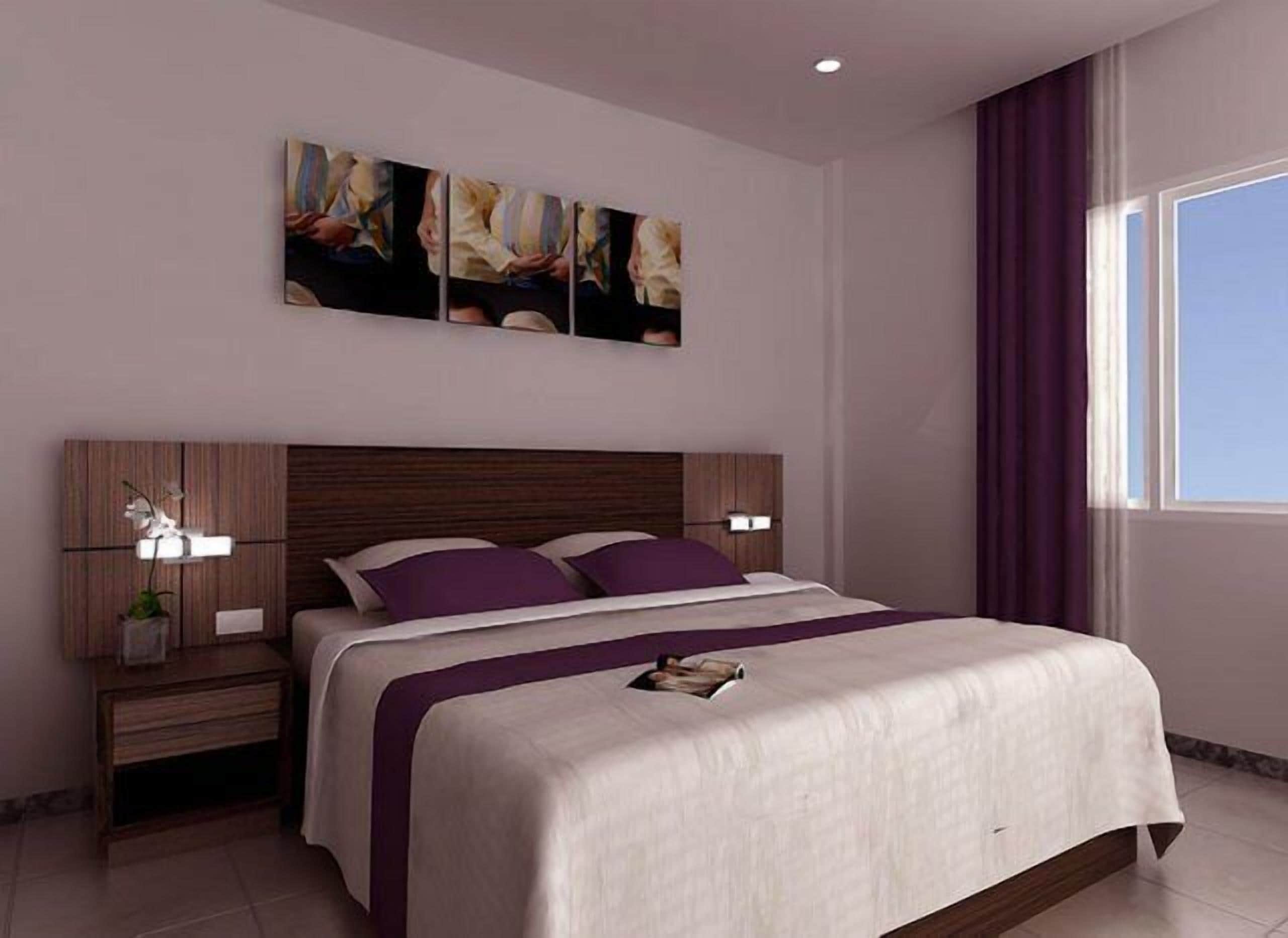 A modern bedroom with a wooden headboard, a large bed with a white and purple bedspread, two bedside tables with lamps, and a window with purple curtains.