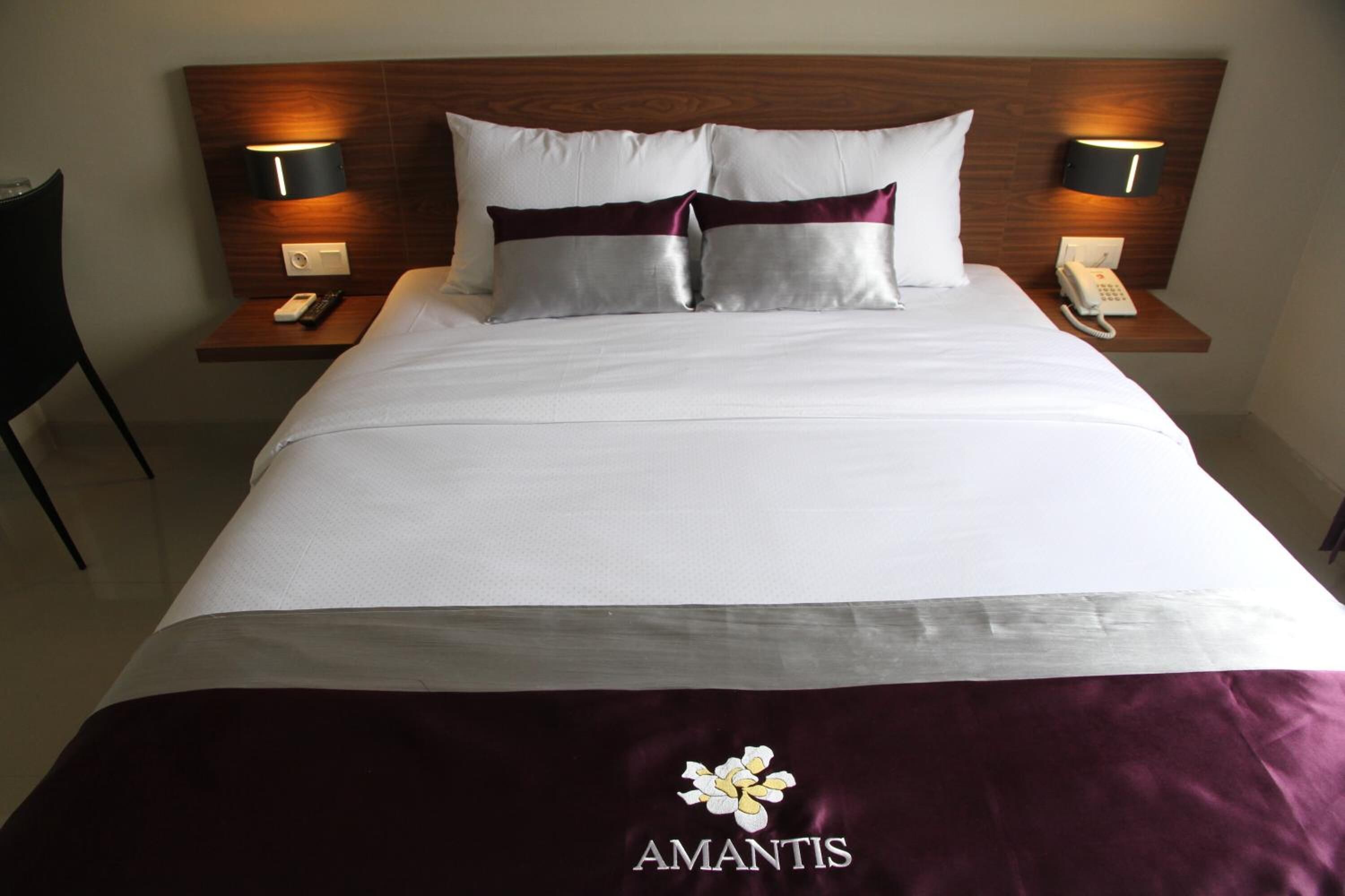 A neatly made bed with white and maroon bedding, two pillows, and a bedspread with the 'Amantis' logo.