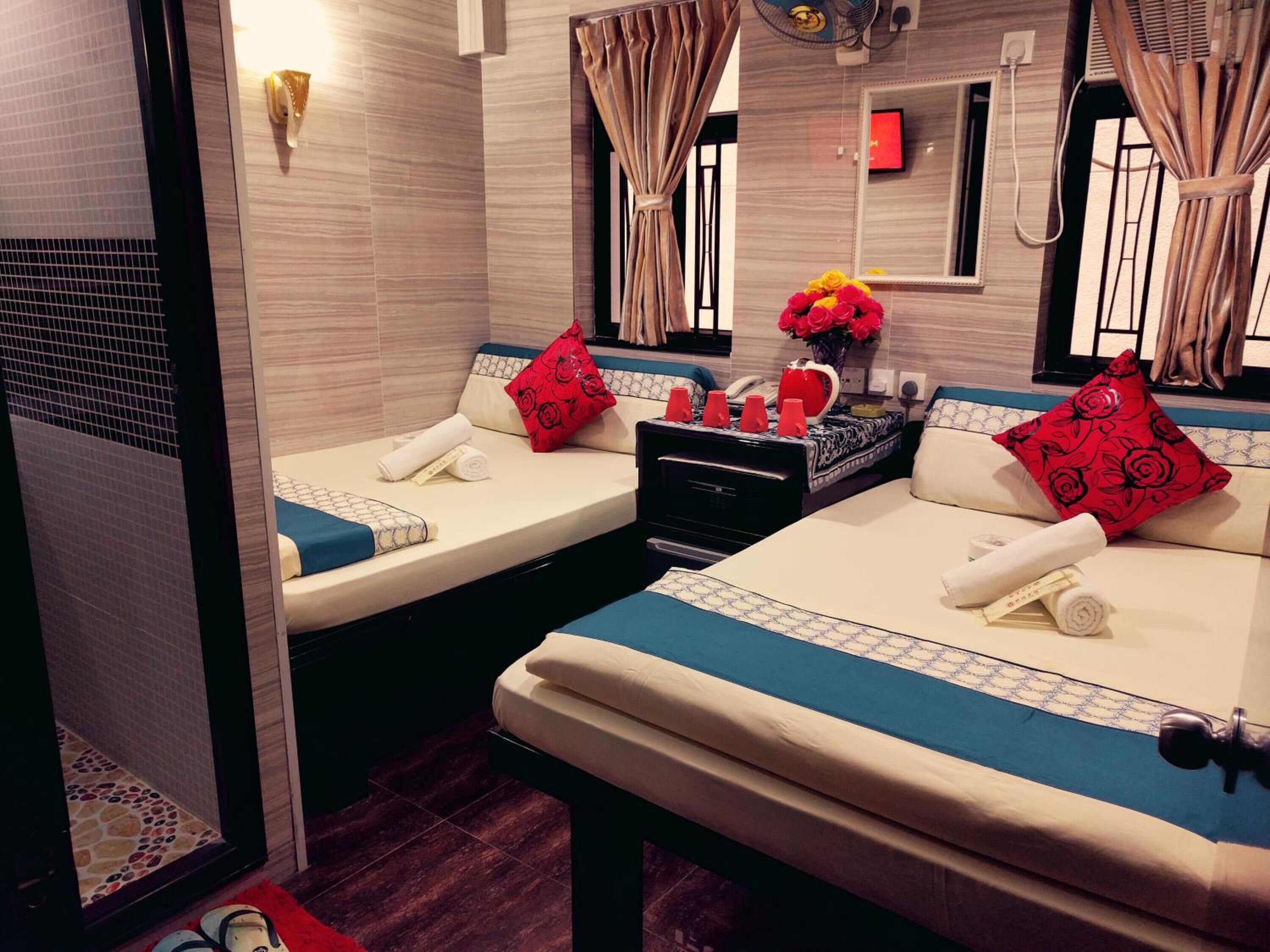 A hotel room with two beds, each with a blue and white patterned bedspread, red floral pillows, and a small table with red cups and a vase of flowers.