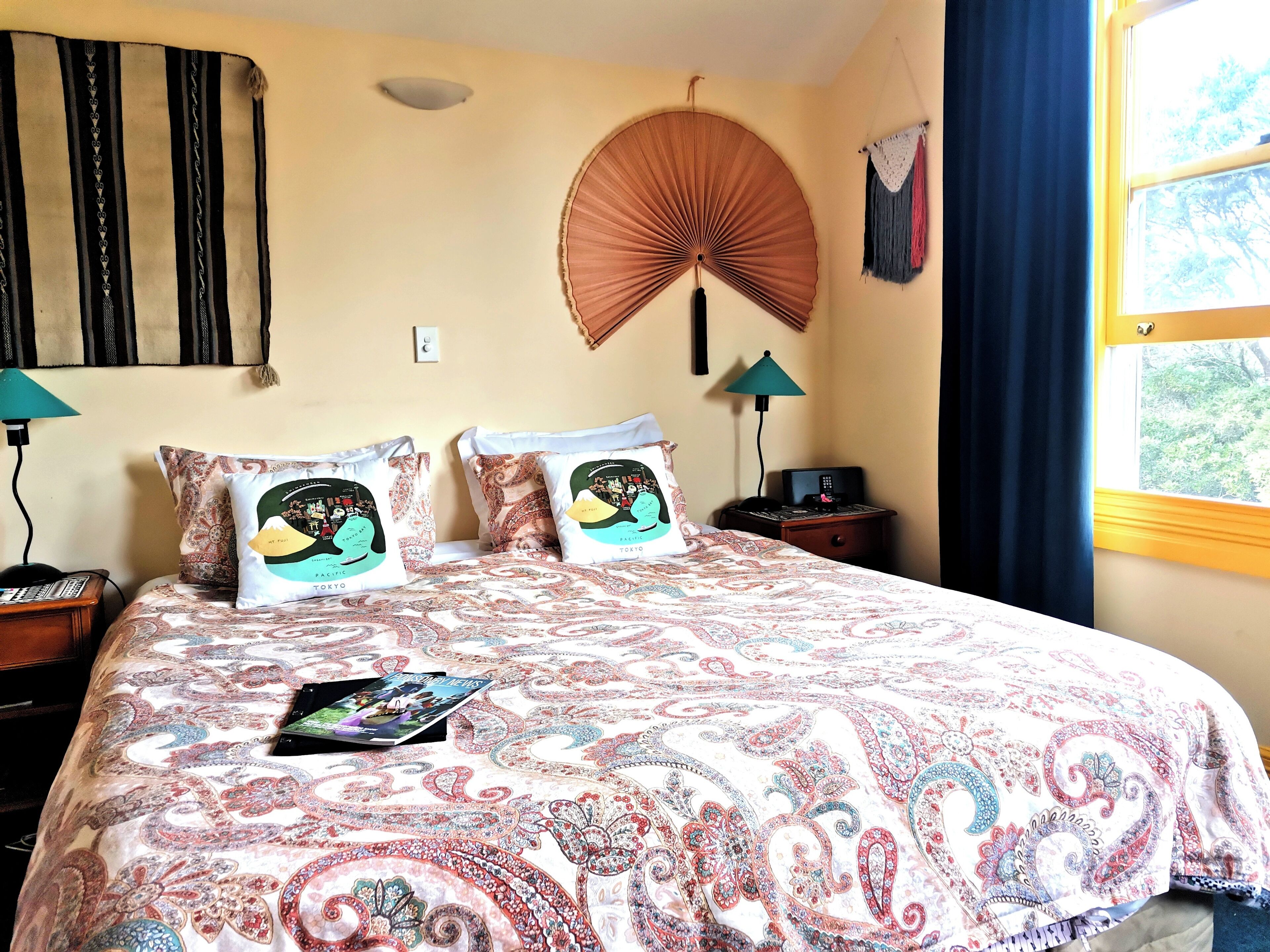 A hotel room with a large bed, two bedside tables, a fan, a lamp, and a window with curtains.