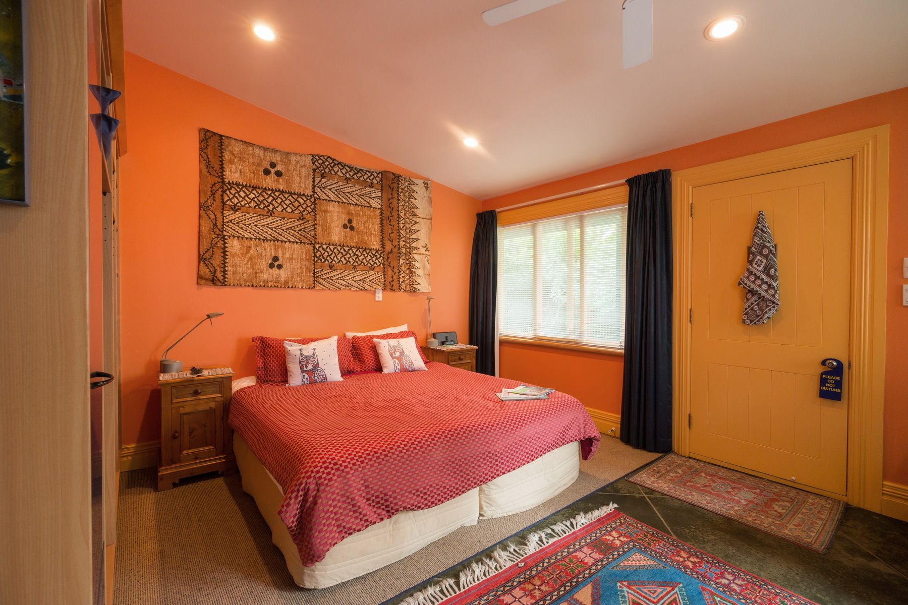 A bedroom with a bed, a nightstand, a wall tapestry, a window with curtains, and a door with a sign.