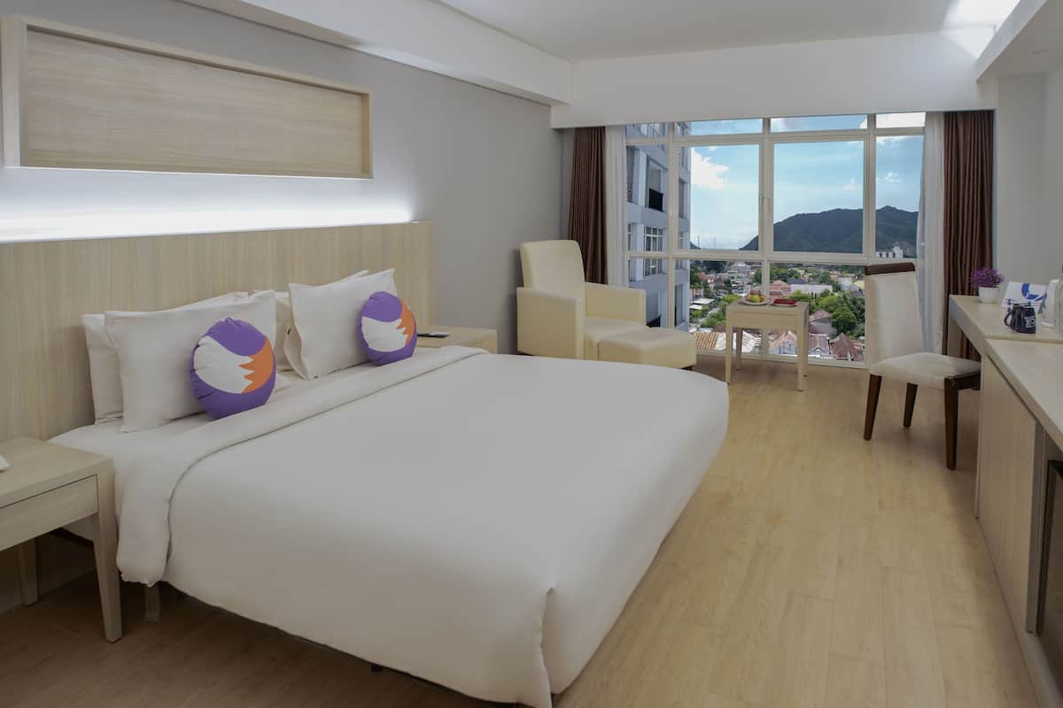 Ein Hotelzimmer mit einem grossen Bett, einem Schreibtisch, einem Stuhl und Blick auf eine Stadtlandschaft durch grosse Fenster.