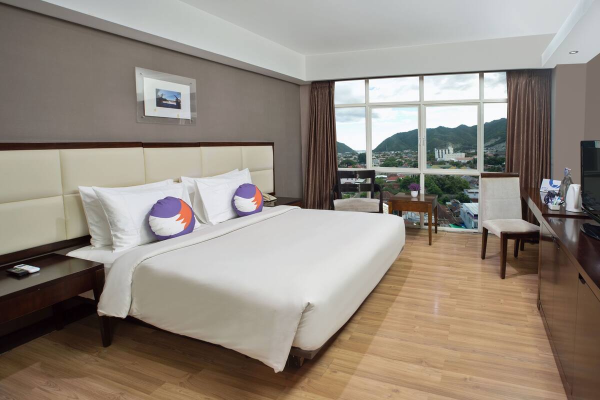 Ein Hotelzimmer mit einem großen Bett, einem Schreibtisch, einem Stuhl und einem Fernseher. Ein Fenster bietet Blick auf Berge und eine Stadt.