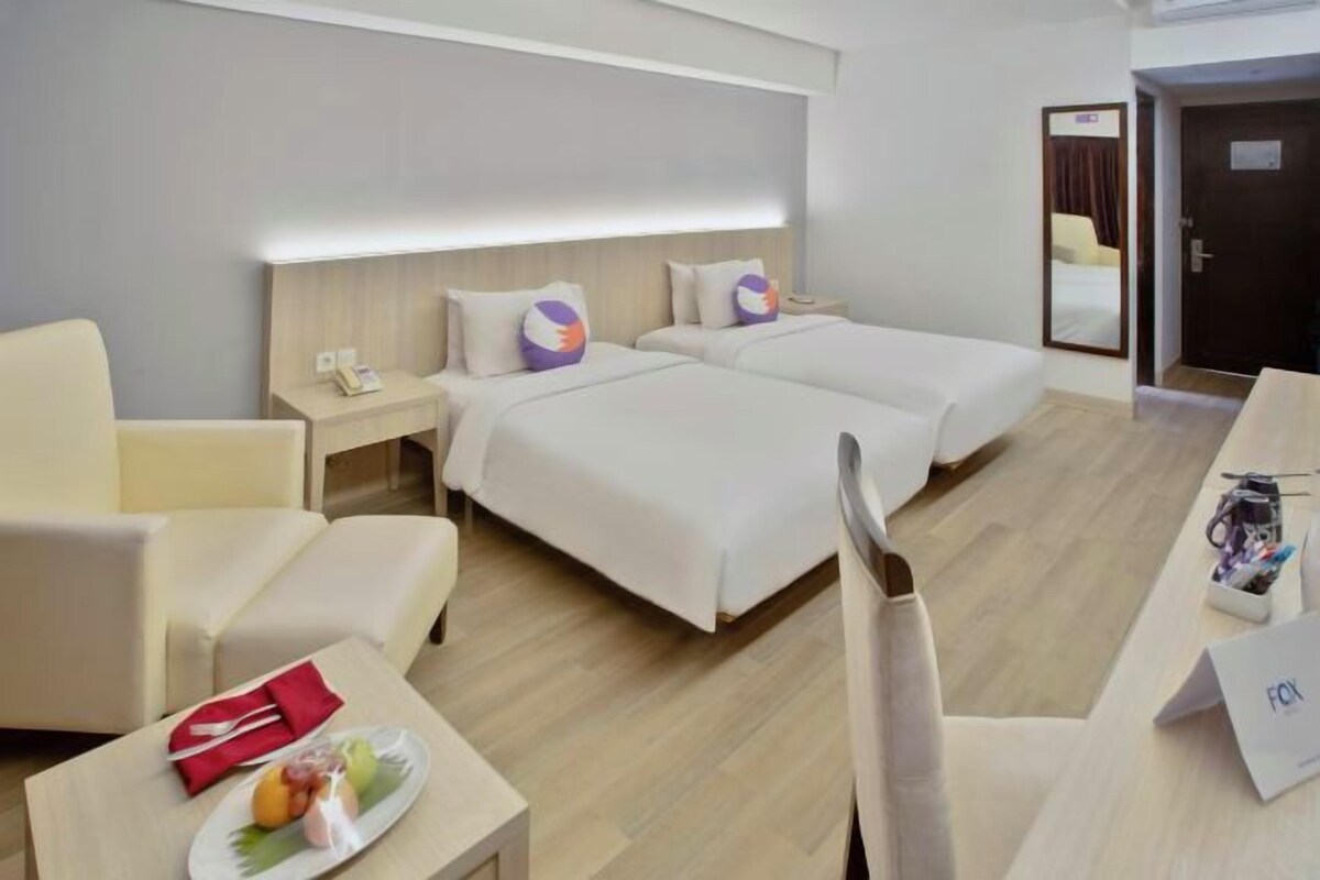 A hotel room with two beds, a desk, a chair, and a small table with a plate of fruit.