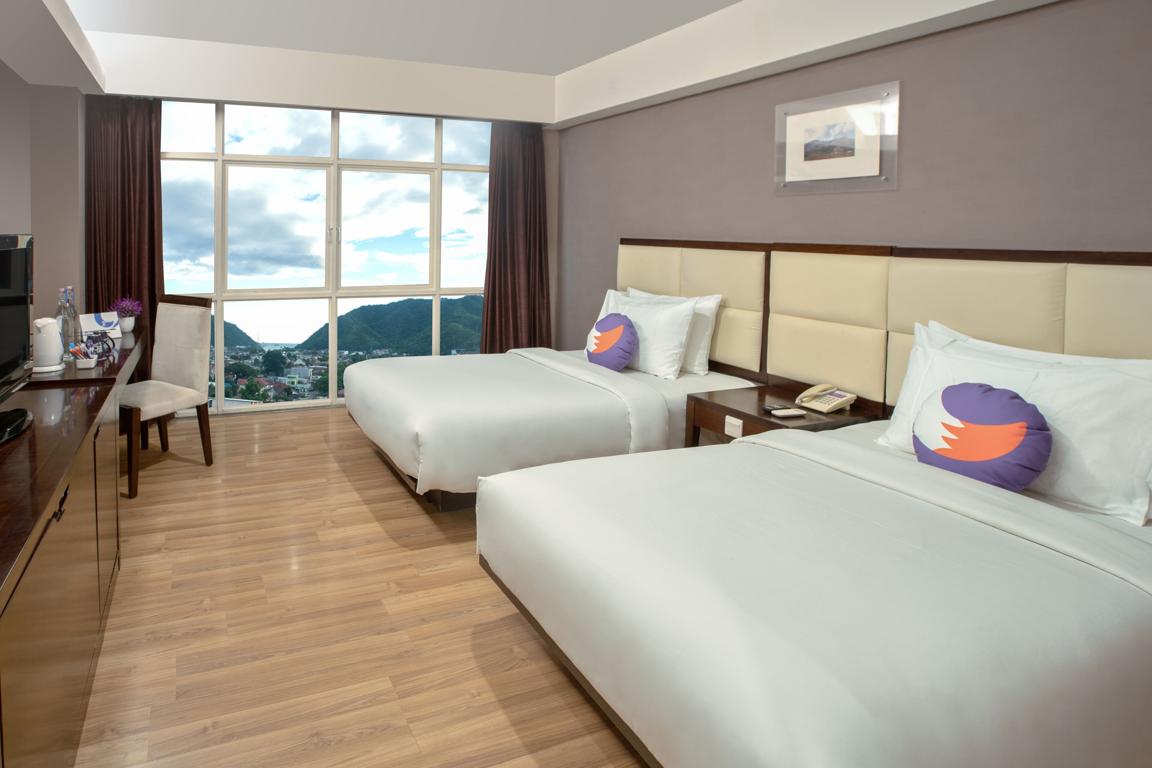 A hotel room with two beds, a desk with a chair, a TV, and a large window offering a city view.