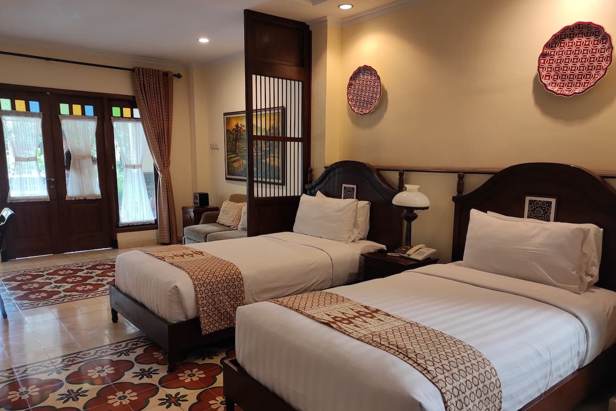 A hotel room with two beds, a patterned floor, a wooden headboard, a nightstand with a lamp, and a painting on the wall.