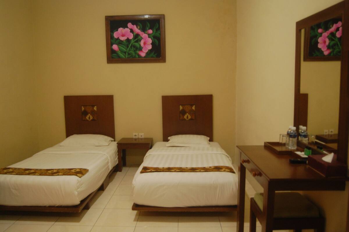 Habitación de hotel con dos camas individuales, cabecera de madera, mesita de noche, escritorio con espejo y una obra de arte floral enmarcada en la pared.