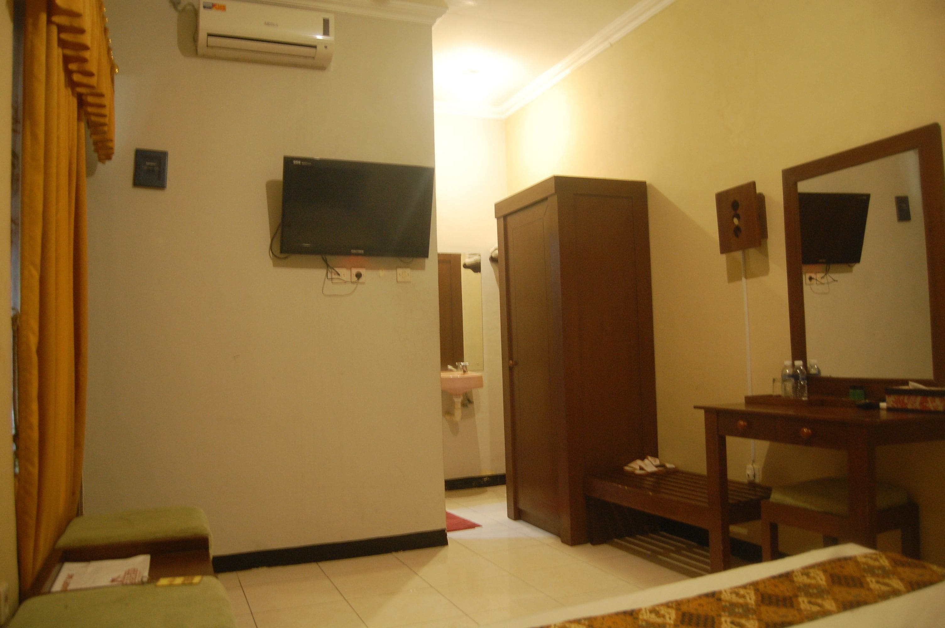 A hotel room with a bed, a wooden wardrobe, a desk with a mirror, a television mounted on the wall, and a bathroom with a sink and mirror.