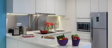 A modern kitchen with white cabinetry, a stainless steel range hood, a built-in oven, a refrigerator, and a central island with a kettle, a toaster, and a bowl of vegetables.