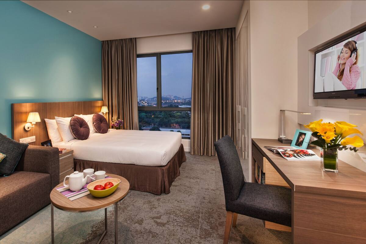 A hotel room with a large bed, a desk, a chair, a small table with a bowl of fruit, and a TV on the wall.