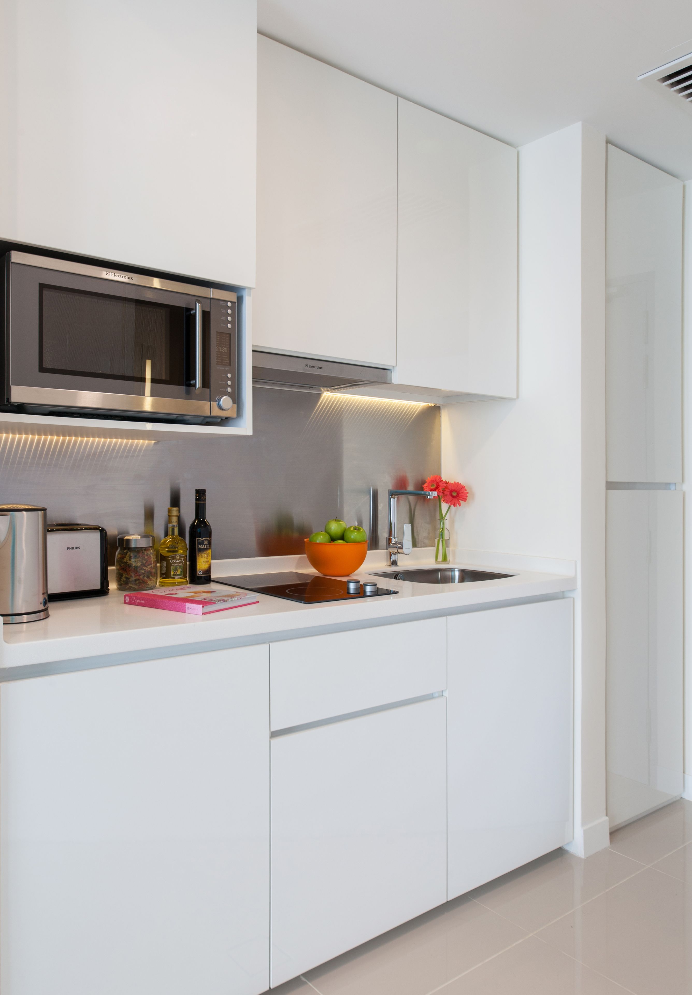 Una cocina moderna con armarios blancos, un microondas de acero inoxidable, un bol de fruta y un florero con una flor.