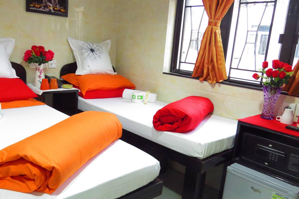 A hotel room with two beds, each with white linens and orange and red pillows. There are two vases with red roses on the nightstands, and a window with curtains.