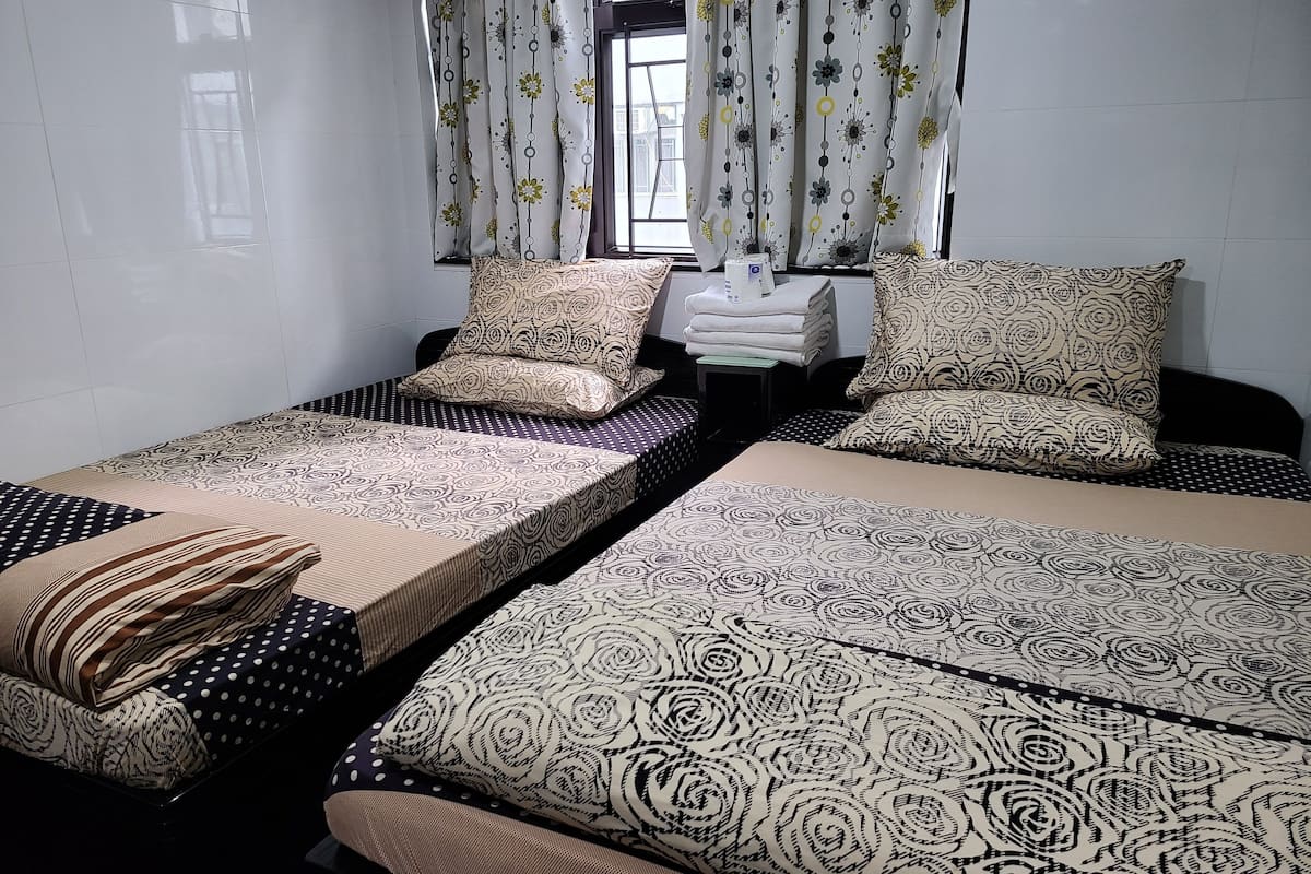 A room with two beds, each with patterned bedding and pillows. There is a window with curtains, and a small table with towels.
